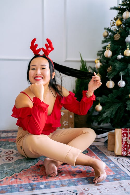 红色意大利面条皮带顶部坐在圣诞树旁边的地板上的女人 · 免费素材图片