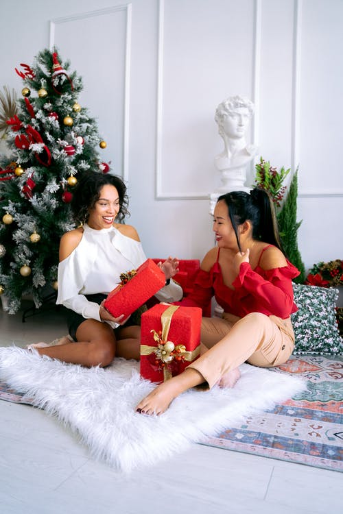 2名妇女坐在绿色圣诞树旁的白色毛皮纺织品上 · 免费素材图片