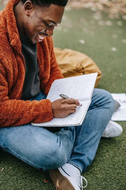 穿着棕色大衣和蓝色牛仔牛仔裤，坐在绿草田间看书的人 · 免费素材图片