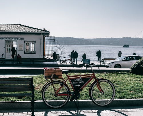 红色城市自行车停在水体附近的棕色木制长椅旁边 · 免费素材图片