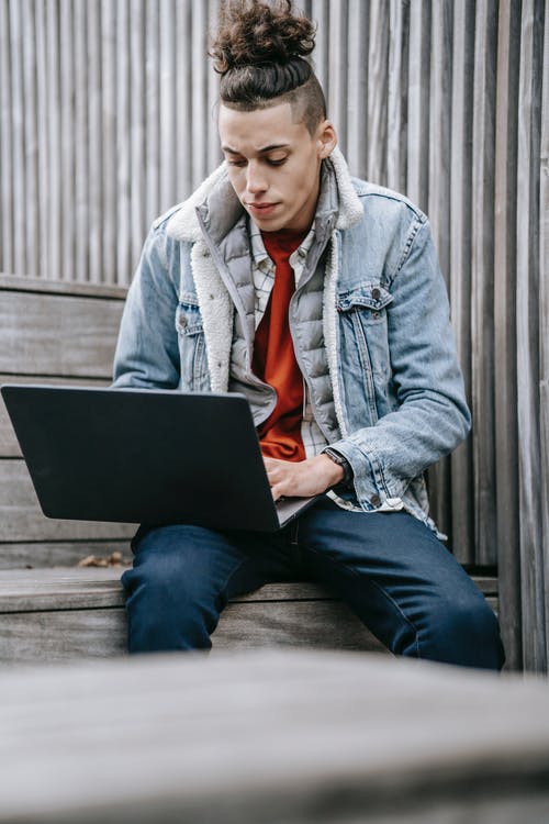 蓝色牛仔夹克和黑色的裤子，坐在黑色的笔记本上的棕色木凳上的男人 · 免费素材图片