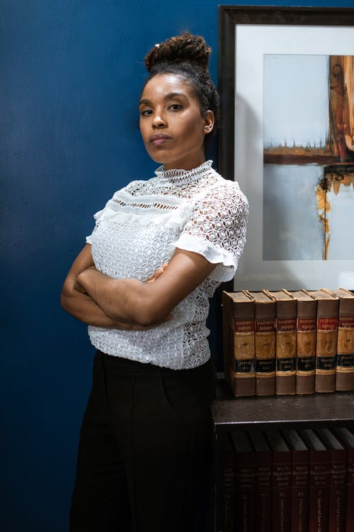 白色和黑色的花衬衫和黑色的裙子，站在棕色木书架旁边的女人 · 免费素材图片