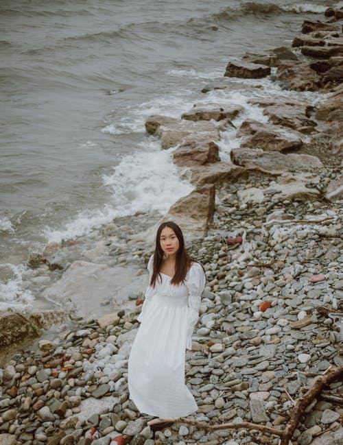 白色礼服站在多岩石的海岸上的女人 · 免费素材图片