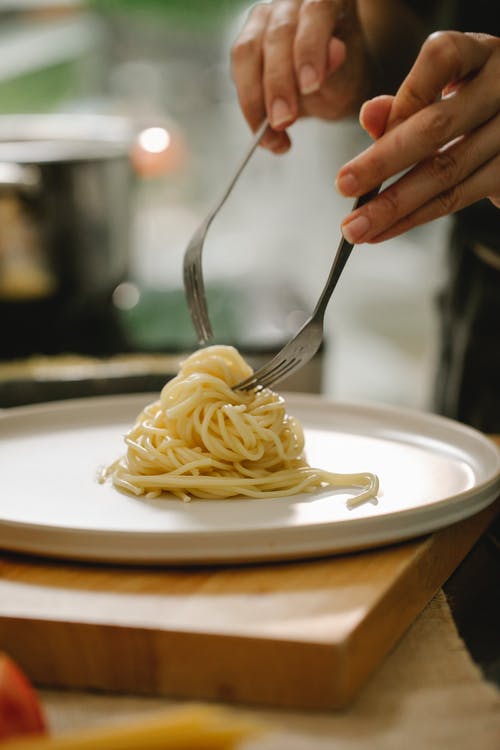 在白色陶瓷板上持有意大利面食的不锈钢叉的人 · 免费素材图片