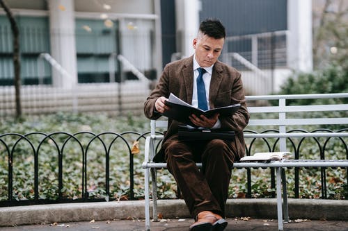 黑色西装的男人坐在白色的金属长凳阅读书上 · 免费素材图片