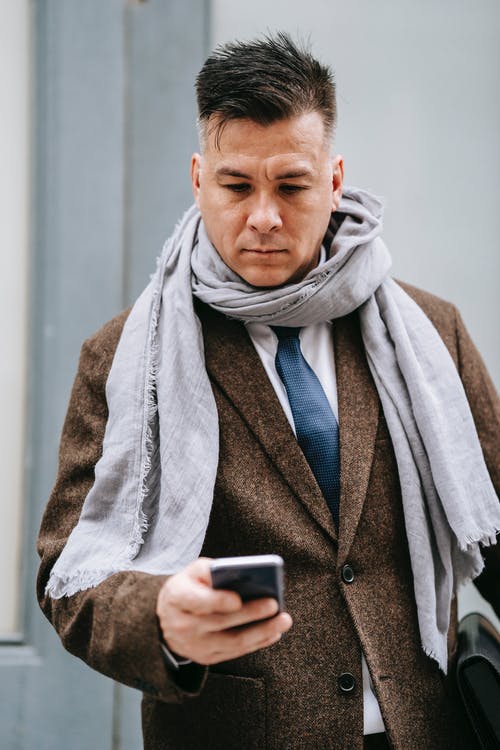 灰色的围巾和棕色的外套，拿着黑色的智能手机的人 · 免费素材图片