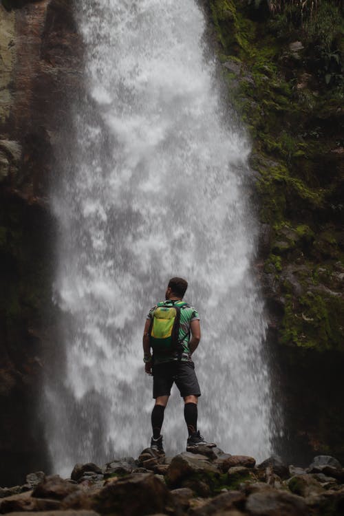 穿着绿色衬衫和黑色短裤，背包里站在瀑布前的人 · 免费素材图片