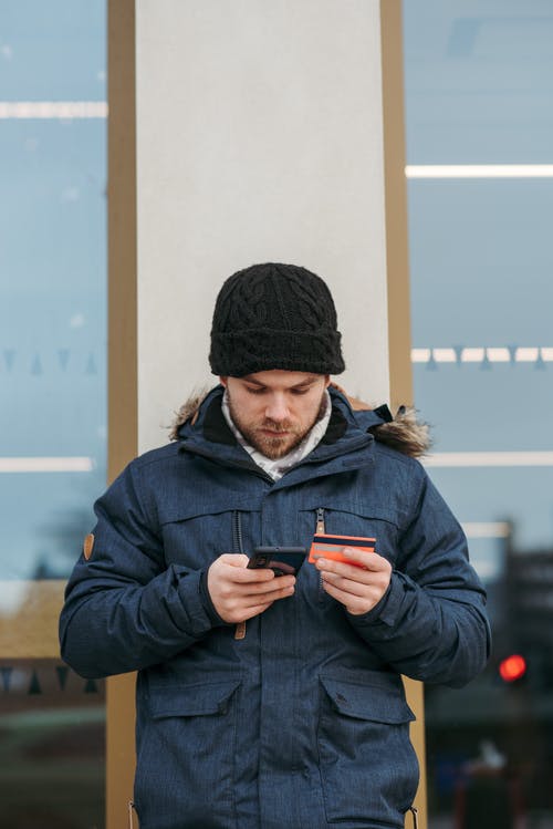 黑色针织帽和黑色外套拿着黑色智能手机的人 · 免费素材图片