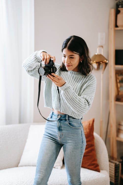 拿着黑色的数码单反相机的白色毛衣和蓝色牛仔牛仔裤的女人 · 免费素材图片
