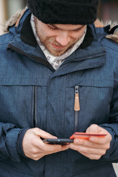 蓝色拉链夹克拿着黑色的智能手机的人 · 免费素材图片