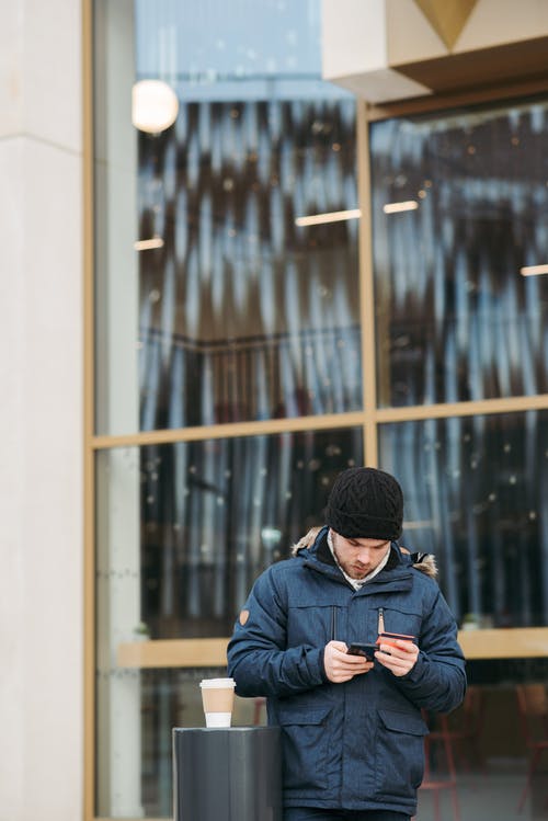 黑色的针织帽和蓝色外套拿着黑色的智能手机的人 · 免费素材图片