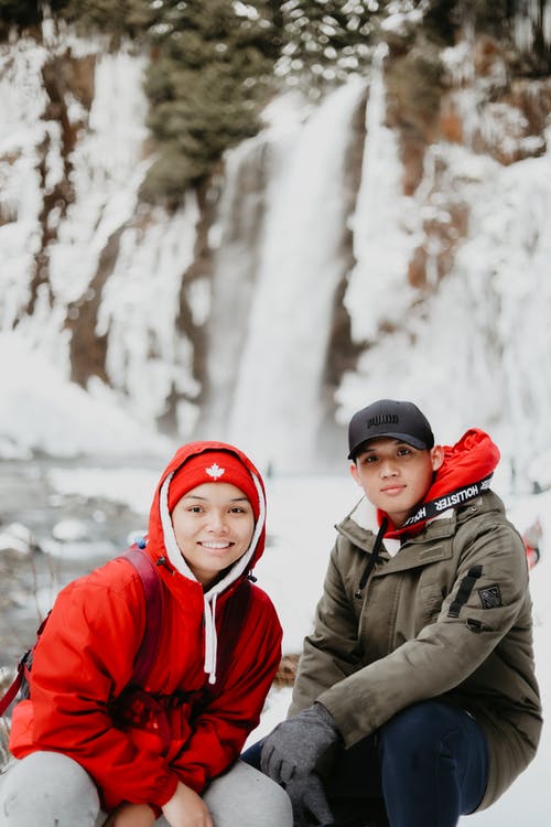 微笑在红色针织帽和灰色夹克站在雪覆盖的树木附近的女人 · 免费素材图片