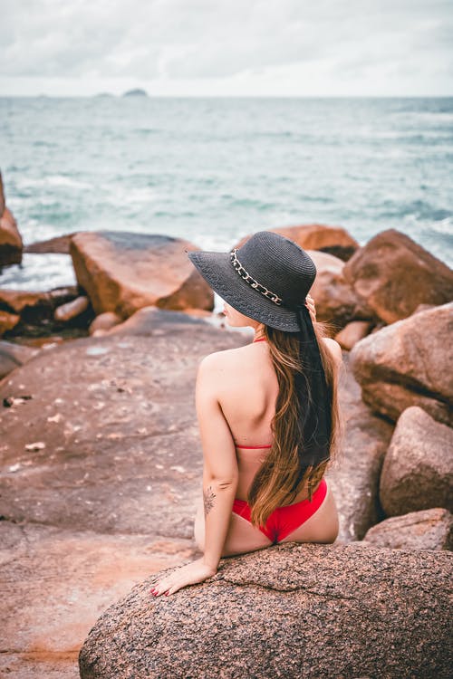 穿着黑色比基尼坐在海边岩石上的红色比基尼的女人 · 免费素材图片