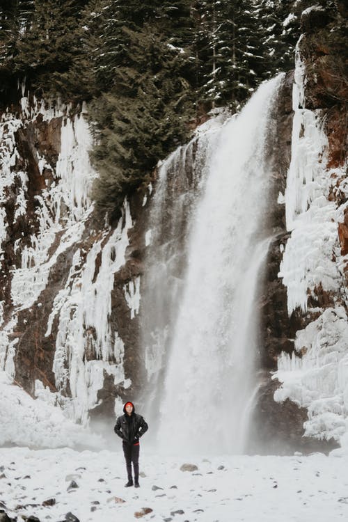 黑夹克和黑裤子的人站在瀑布附近的积雪覆盖的地面上 · 免费素材图片