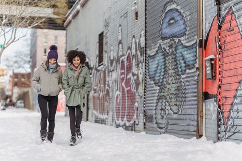 2名妇女站在白雪皑皑的棕色房子附近的积雪的地面上 · 免费素材图片