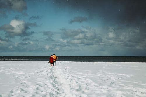 橙色夹克的人在蓝色和白色的晴朗多云的天空下的雪覆盖的田野上行走 · 免费素材图片