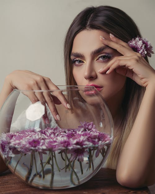在盛满紫色花朵的玻璃碗旁摆姿势的女人 · 免费素材图片
