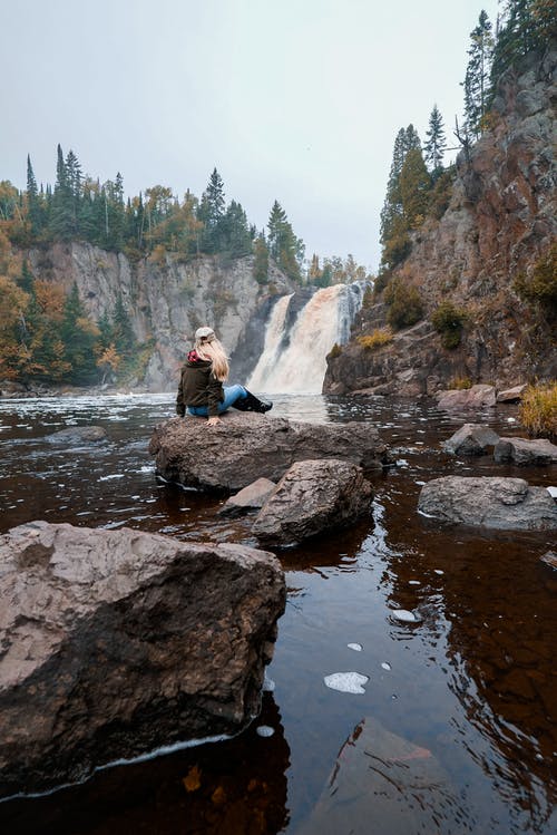 女人坐在瀑布前的岩石上 · 免费素材图片