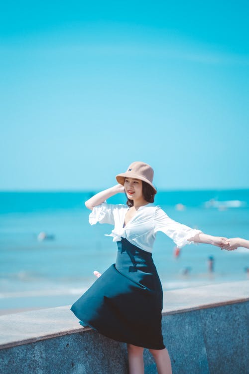 白色的长袖衬衫和黑色的裙子，戴着白帽子站在海边的女人 · 免费素材图片