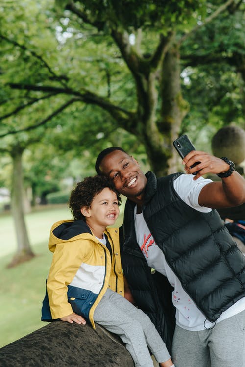 快乐黑人父亲与儿子在智能手机上采取自拍照户外 · 免费素材图片