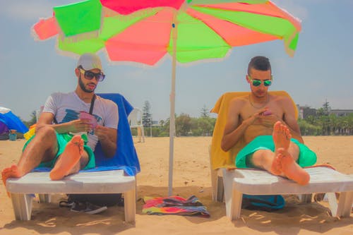 2名男子坐在白色的沙滩躺椅之间，中间有绿色和粉红色的露台伞 · 免费素材图片
