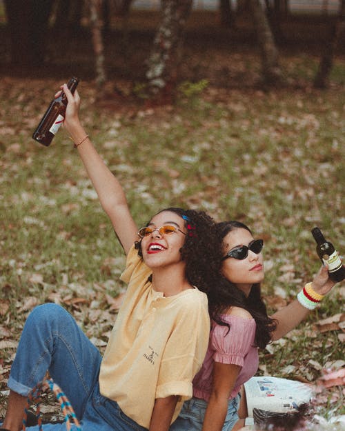 拿着瓶的两个微笑的妇女坐在绿草领域 · 免费素材图片