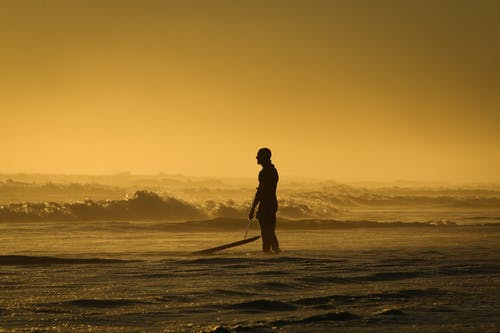 在日落时按住他的冲浪板站在海边的人 · 免费素材图片