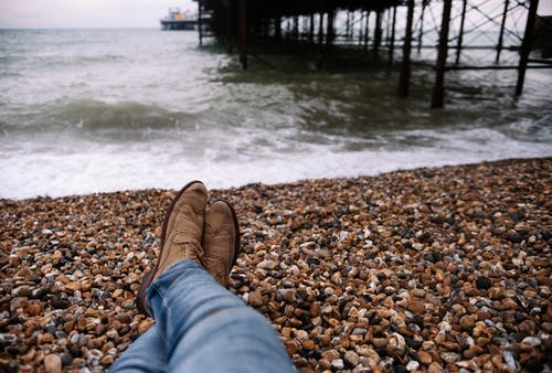 穿着蓝色牛仔长裤和棕色靴子坐在水体前的棕色和黑色石头上的人 · 免费素材图片