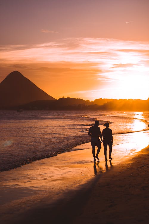 两人在日落时在海滩上行走的剪影 · 免费素材图片