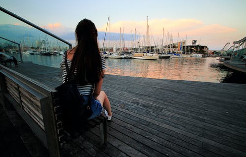 黑色和白色的条纹勺脖子衬衫的女人在白天坐在船坞的棕色木凳上 · 免费素材图片
