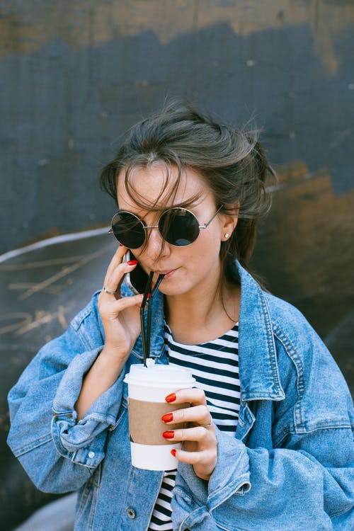 蓝色牛仔夹克的女人拿着一次性杯子和电话交谈 · 免费素材图片