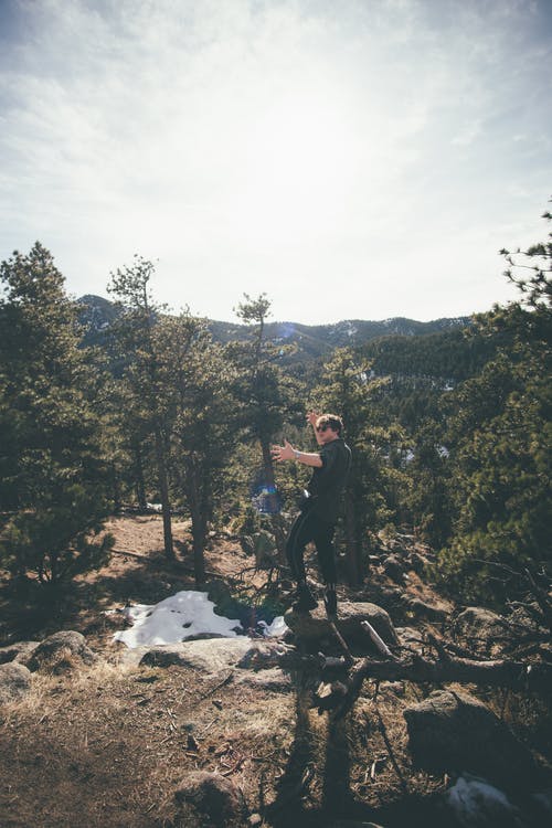 穿黑夹克的男人站在白云下绿树附近的岩石地面上 · 免费素材图片