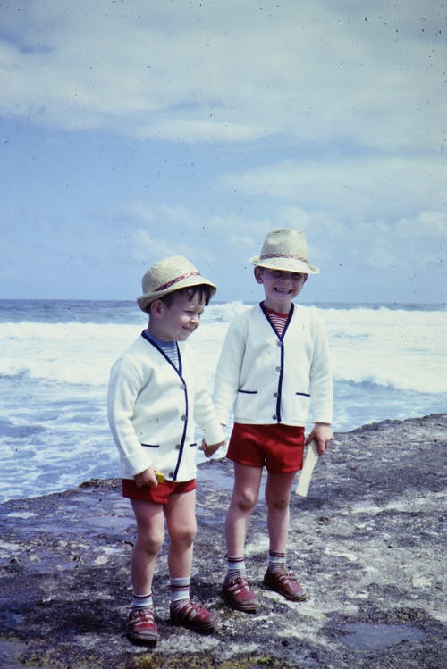 两个男孩穿着白色毛衣和红色短裤站在海滩上 · 免费素材图片
