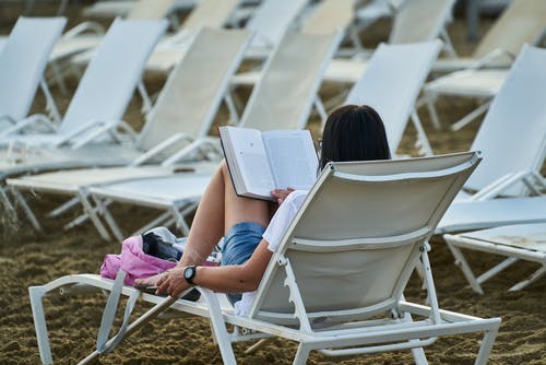 蓝色牛仔短裤的妇女坐在白色折叠椅阅读书 · 免费素材图片