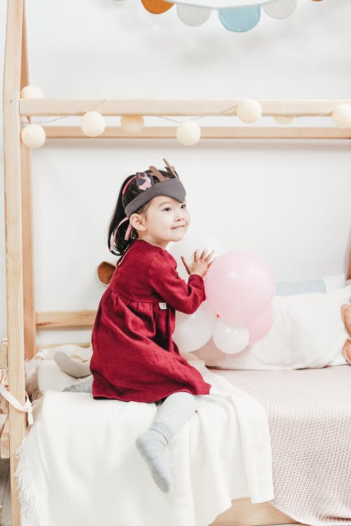 红色长袖穿裙子坐在床上玩气球的女孩 · 免费素材图片