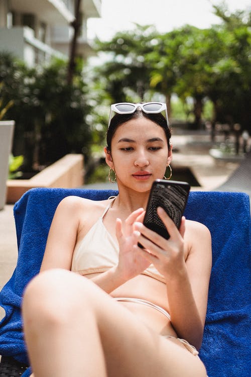 年轻的族裔女人浏览智能手机，而冷在日光浴浴床日光浴浴床 · 免费素材图片