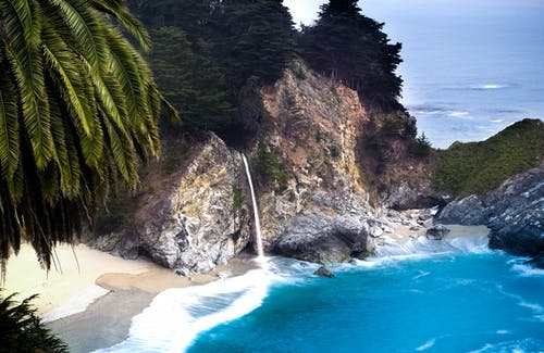 棕色和灰色岩石与绿叶树附近的蓝色大海 · 免费素材图片