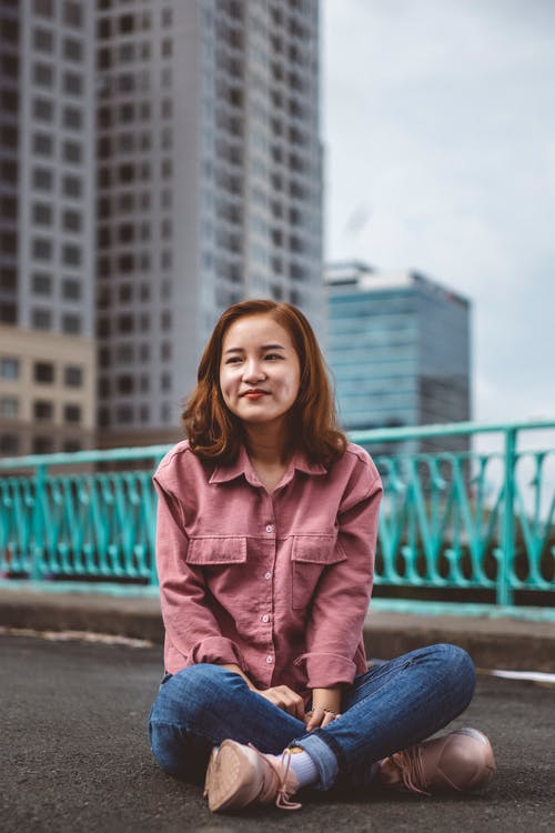 微笑的女人，穿着蓝色牛仔裤和粉红色的衬衫，坐在水泥地上的选择性焦点照片 · 免费素材图片