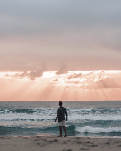 日落时暴风雨海洋附近的海滩上无法识别的男性旅行者 · 免费素材图片