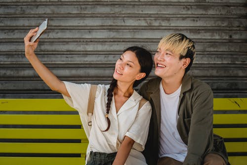 微笑亚洲夫妇在长凳上的智能手机上采取自拍照 · 免费素材图片
