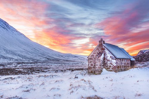 黄色蓝色和橙色日落下的白雪覆盖的山脉附近的白雪覆盖领域的棕色房子 · 免费素材图片