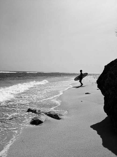 人站在海边拿着冲浪板的灰度照片 · 免费素材图片