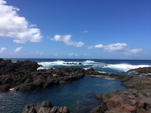 棕色岩石在海洋旁边，气泡波在蓝蓝的天空下 · 免费素材图片