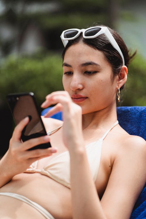 集中躺在泳装日光浴浴床上冲浪智能手机的年轻族裔小姐 · 免费素材图片
