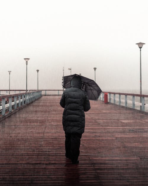 身穿黑色外套和黑色的裤子，拿着伞在木板路上行走的人 · 免费素材图片