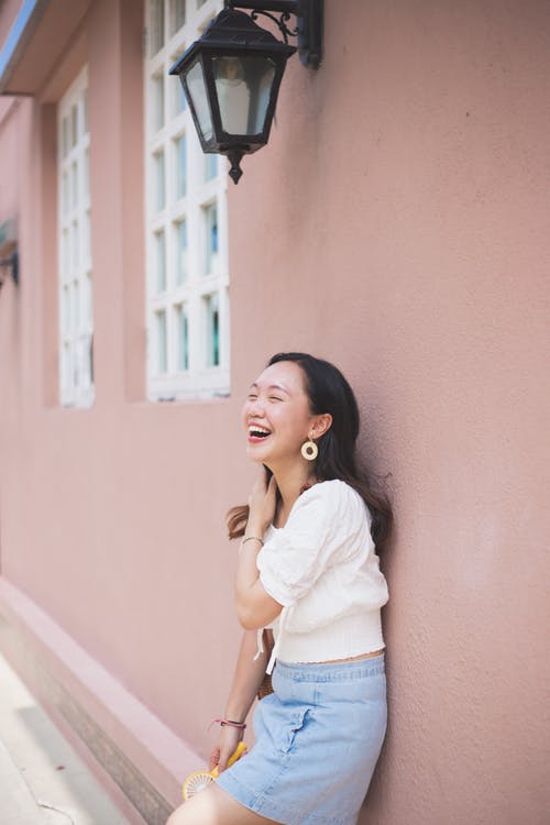 年轻的亚裔女子，同时靠在墙上时开心地笑 · 免费素材图片