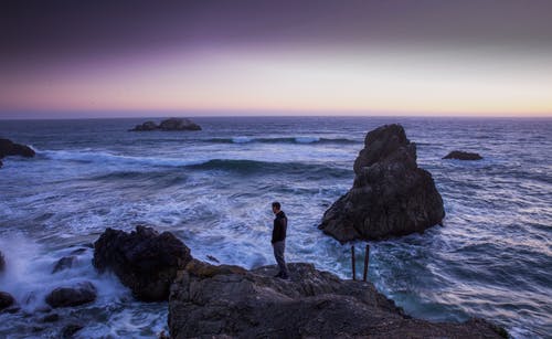 黑色衬衫的男人站在海水之间的岩石上 · 免费素材图片