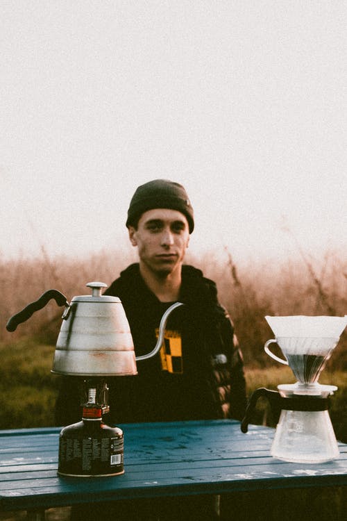 戴着黑夹克的男子站在木制的桌子上的便携式丙烷罐顶部的水壶上凝视着 · 免费素材图片