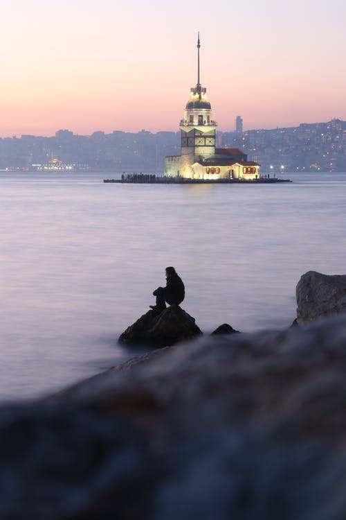 在日落期间坐在水附近的岩石上的人的身影 · 免费素材图片