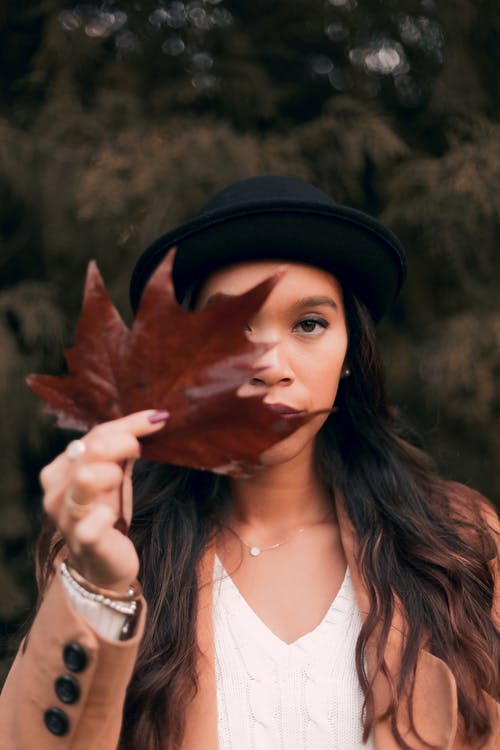 女人拿着棕色的枫叶时穿棕色的外套和黑色的帽子 · 免费素材图片